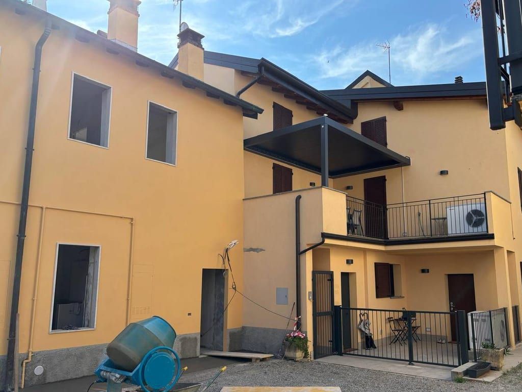 Il cortile a lavori finiti: edifici intonacati di giallo, pergola scura sul terrazzo, balcone con la ringhiera nera e cancello al piano terra
