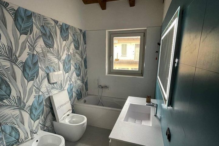 Il bagno finito: vasca sotto la finestra, parete decorata a foglie, mobile lavabo e specchio con luce